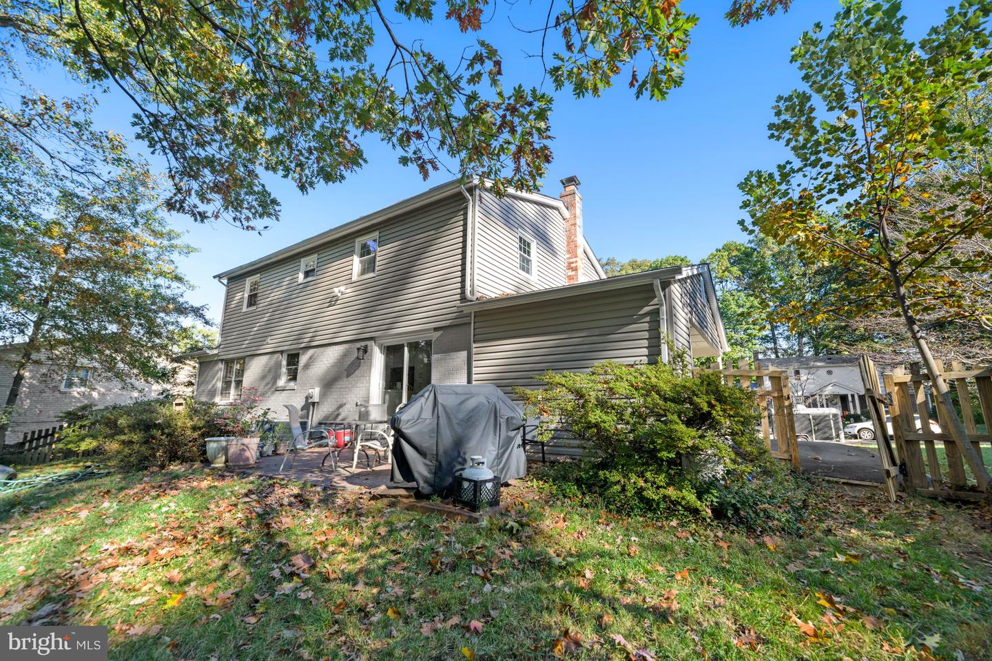 Rear exterior of a two-story gray-sided house with a brick chimney and tree-shaded patio.