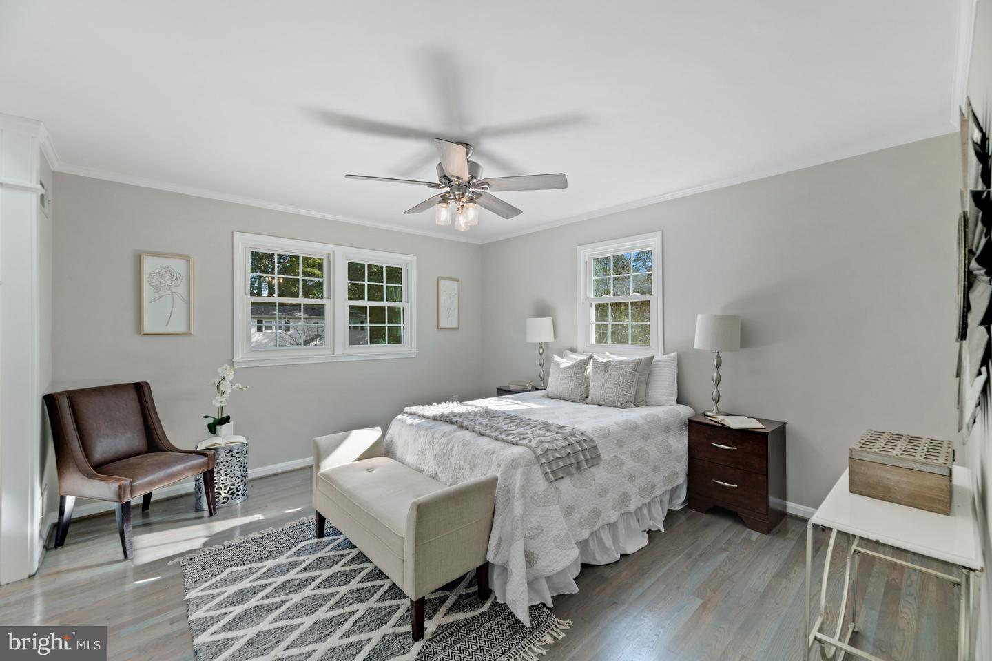 Staged bedroom with a white bed, patterned rug, upholstered bench, and ceiling fan.