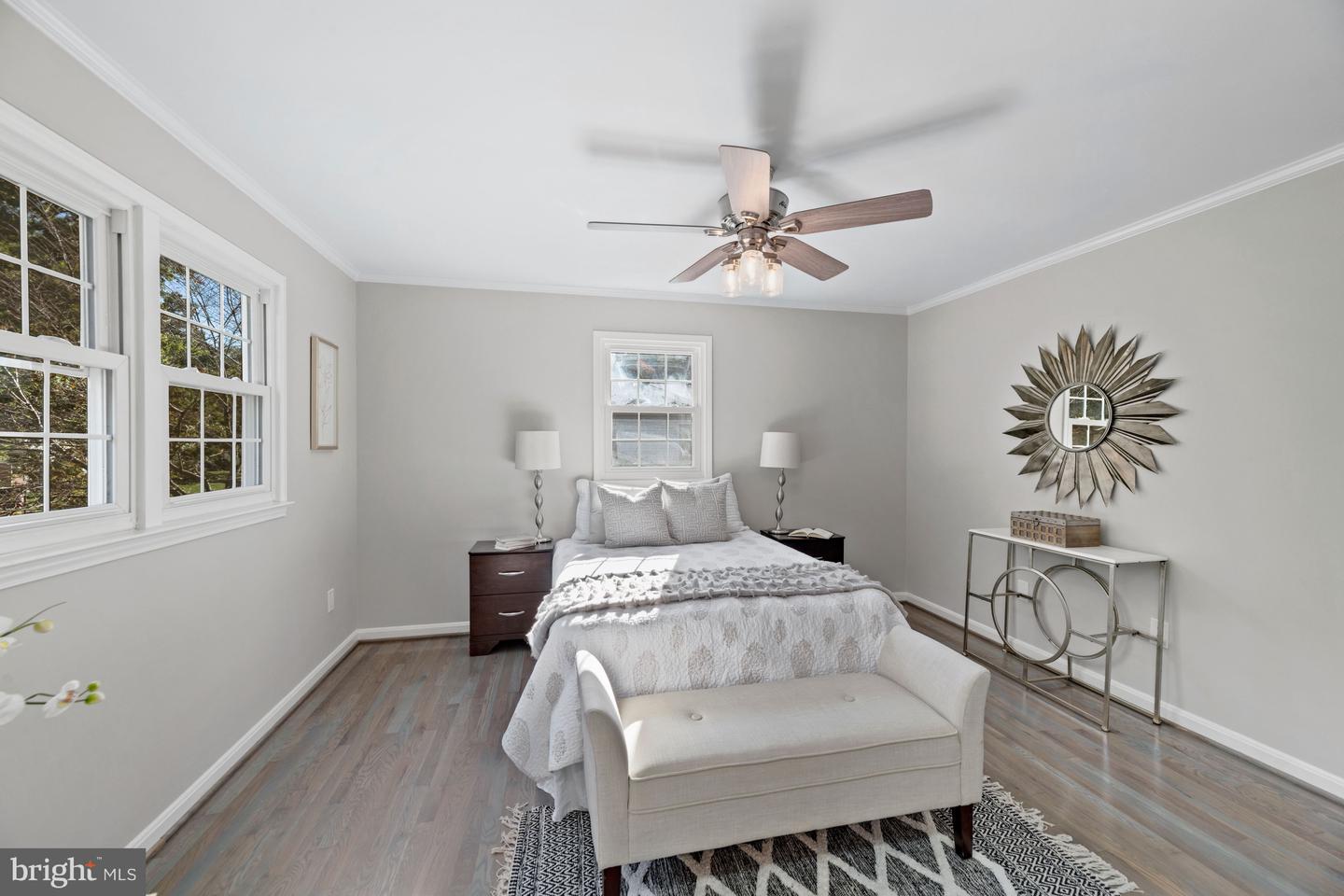 Staged bedroom with a white bed, large windows, ceiling fan, and sunburst mirror.