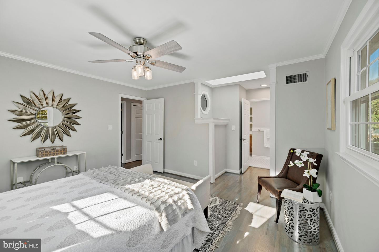 Staged bedroom with a white bed, ceiling fan, sunburst mirror, and large windows.