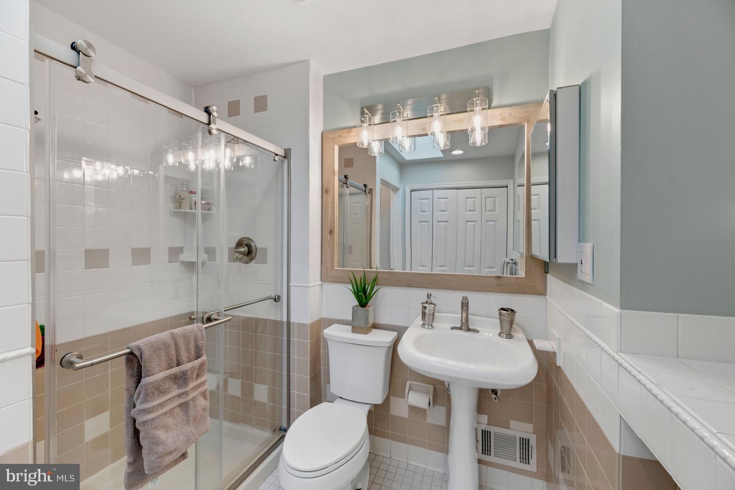 Bathroom with glass shower, beige-and-white tile, pedestal sink, and large wood-framed mirror.
