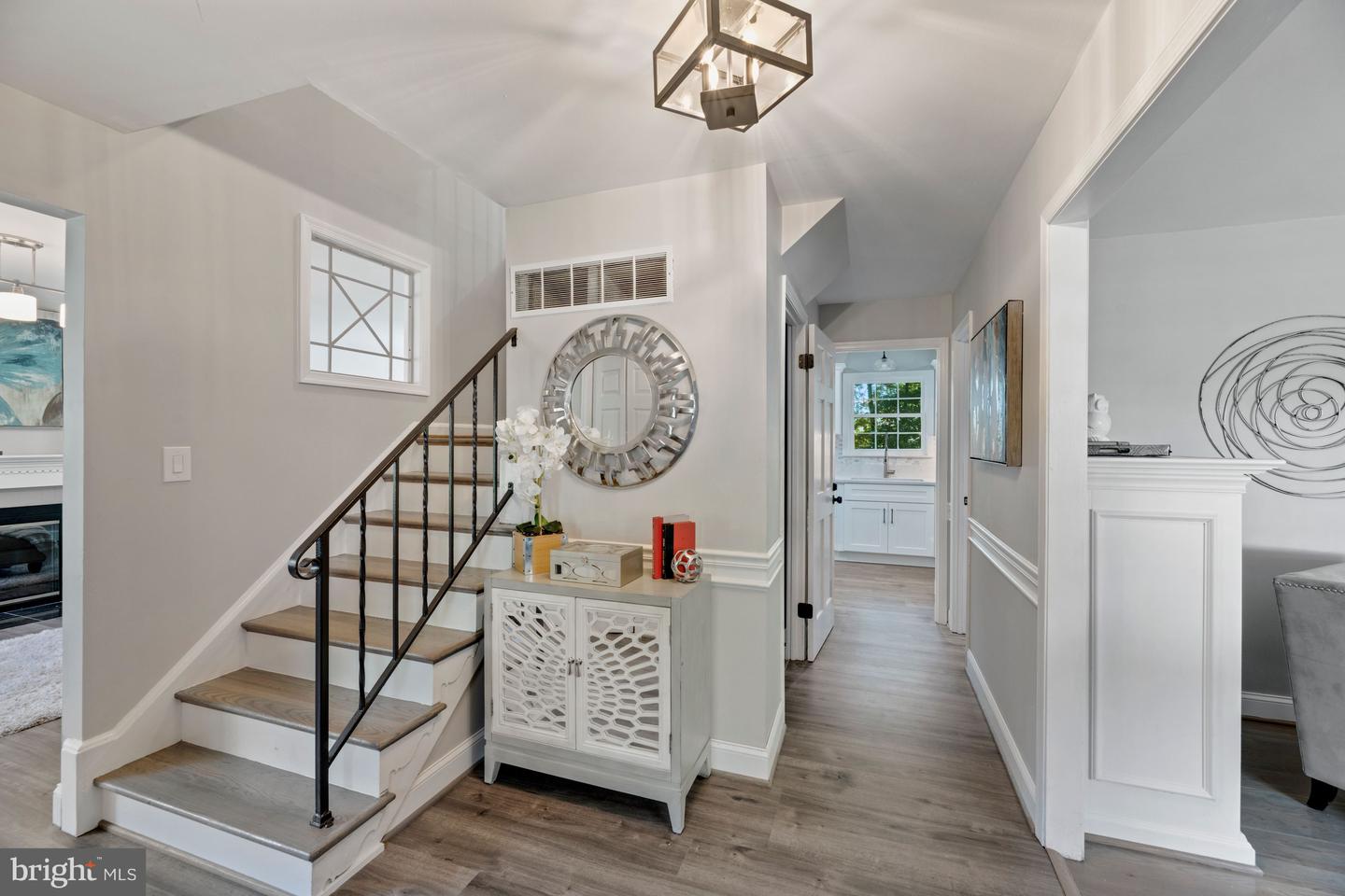 Staged entry hall with a black-railed staircase, round mirror, and view into a white kitchen.