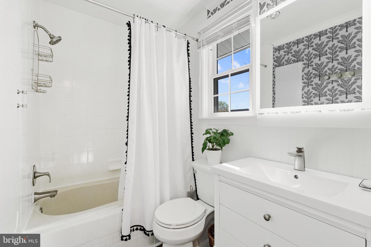 Bright bathroom with a white bathtub, tasseled shower curtain, and vanity beneath a window.