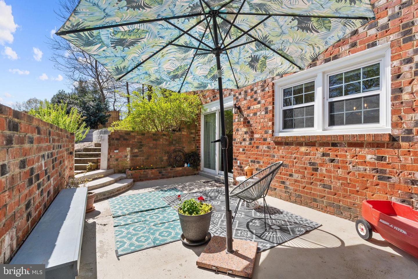 Brick patio with a patterned umbrella, outdoor rugs, and a black wire chair beside the home.