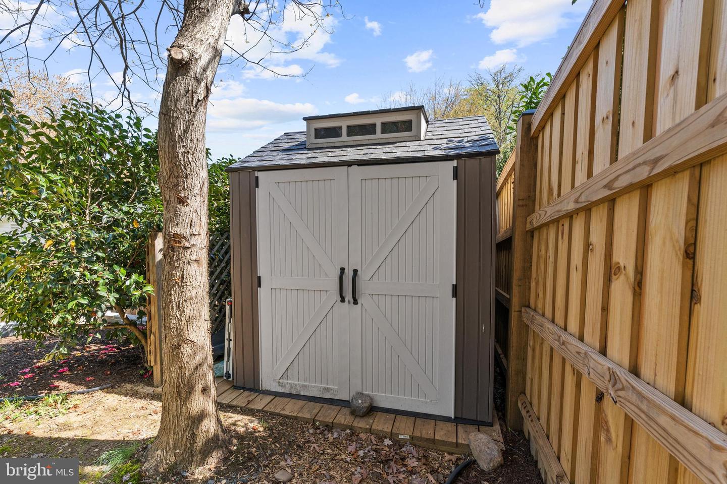 Backyard storage shed with white double doors beside a wooden privacy fence and mature tree.