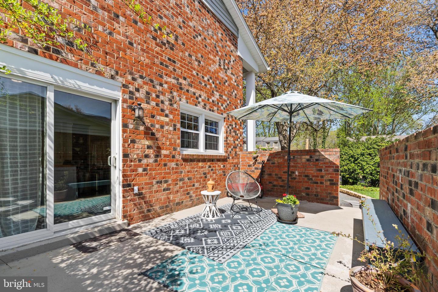 Brick patio with an umbrella, lounge chair, patterned rugs, and potted plants.
