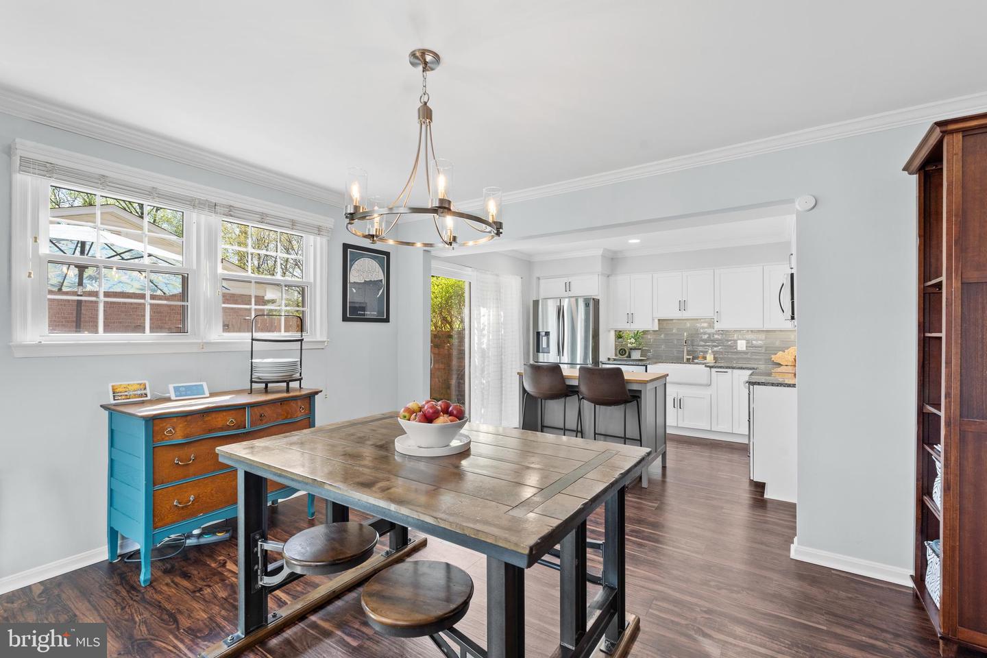 Dining area with a rustic wood table and blue chest, opening to a bright white kitchen with stainless appliances.