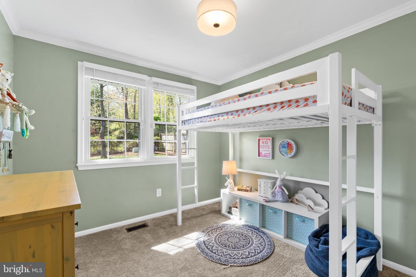 Furnished child’s bedroom with a white loft bed, sage green walls, and windows overlooking a leafy yard.
