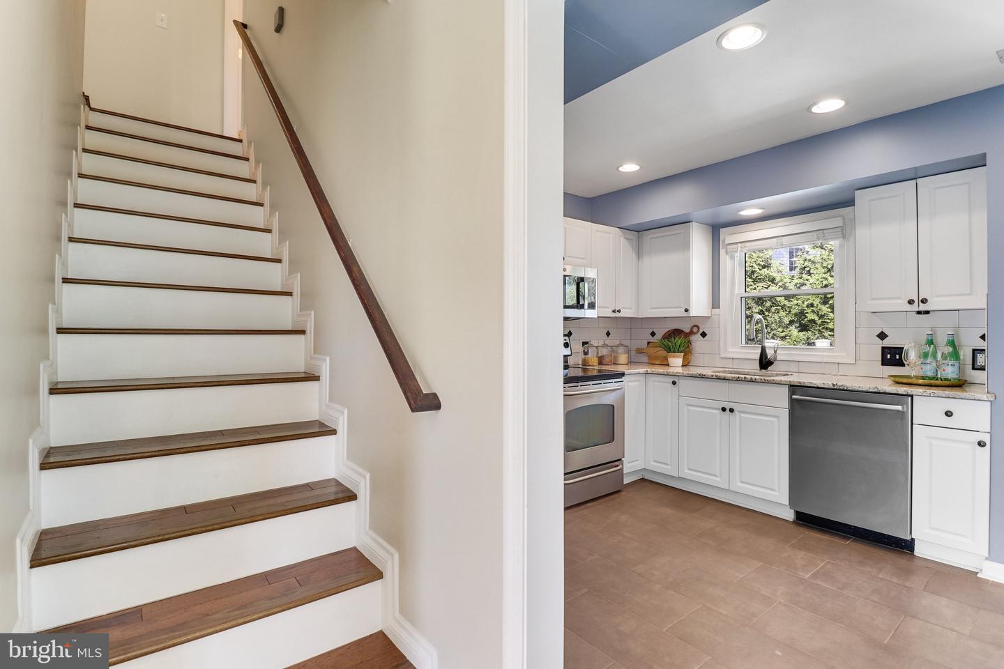 White kitchen with stainless appliances beside a staircase with wood treads and a dark handrail.