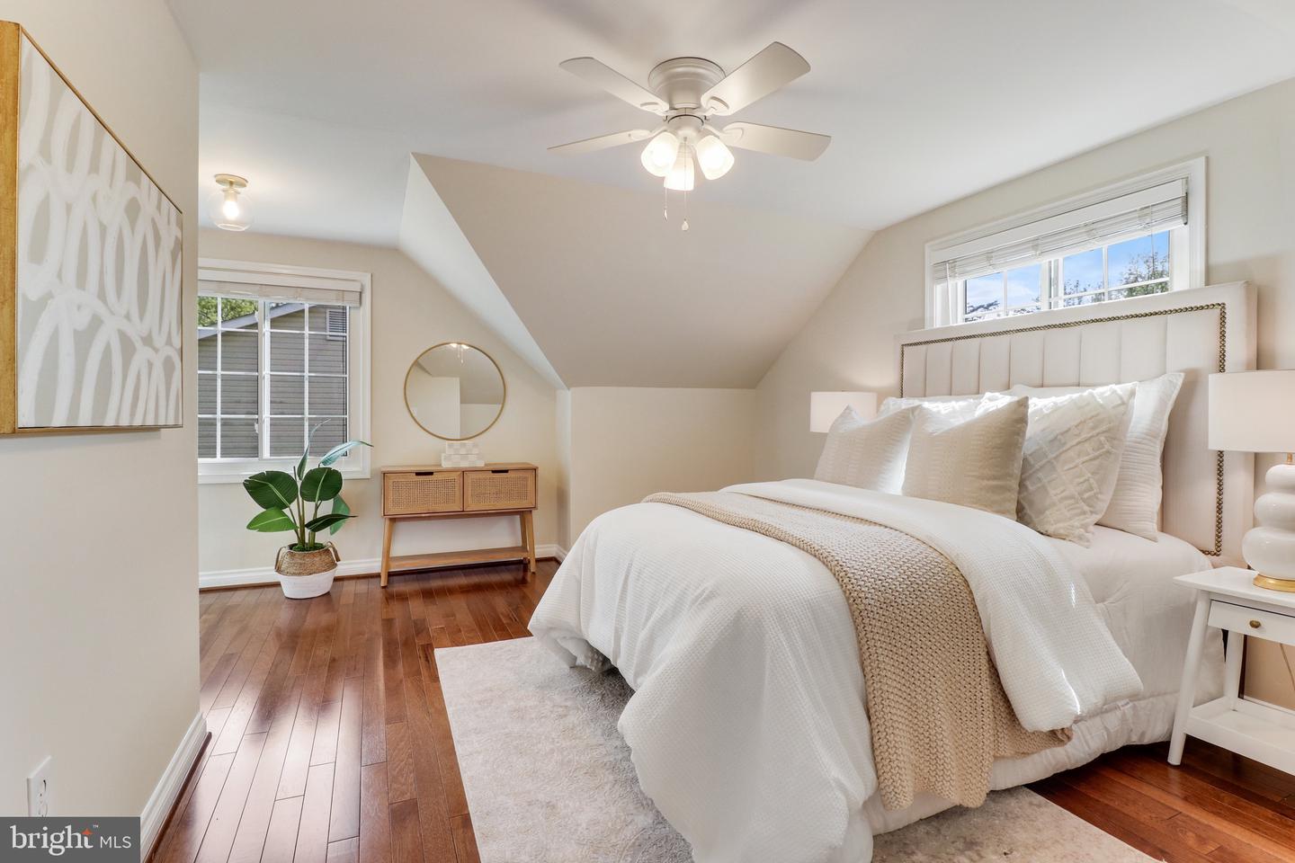 Staged bedroom with a white upholstered bed, warm hardwood floors, and a ceiling fan.