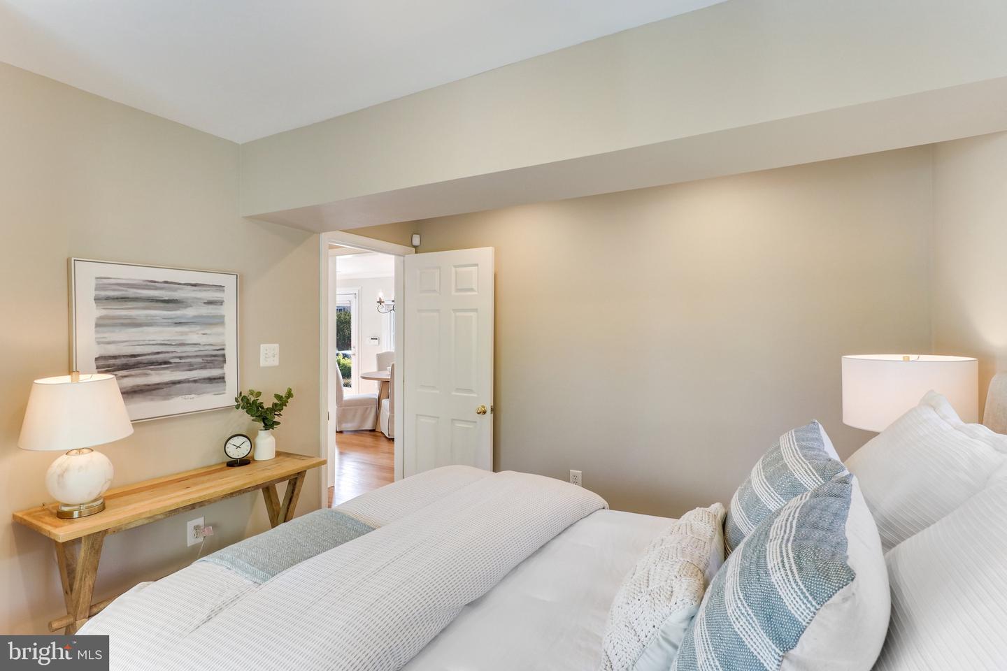 Staged bedroom with layered bedding, abstract wall art, and an open door to a bright adjoining room.