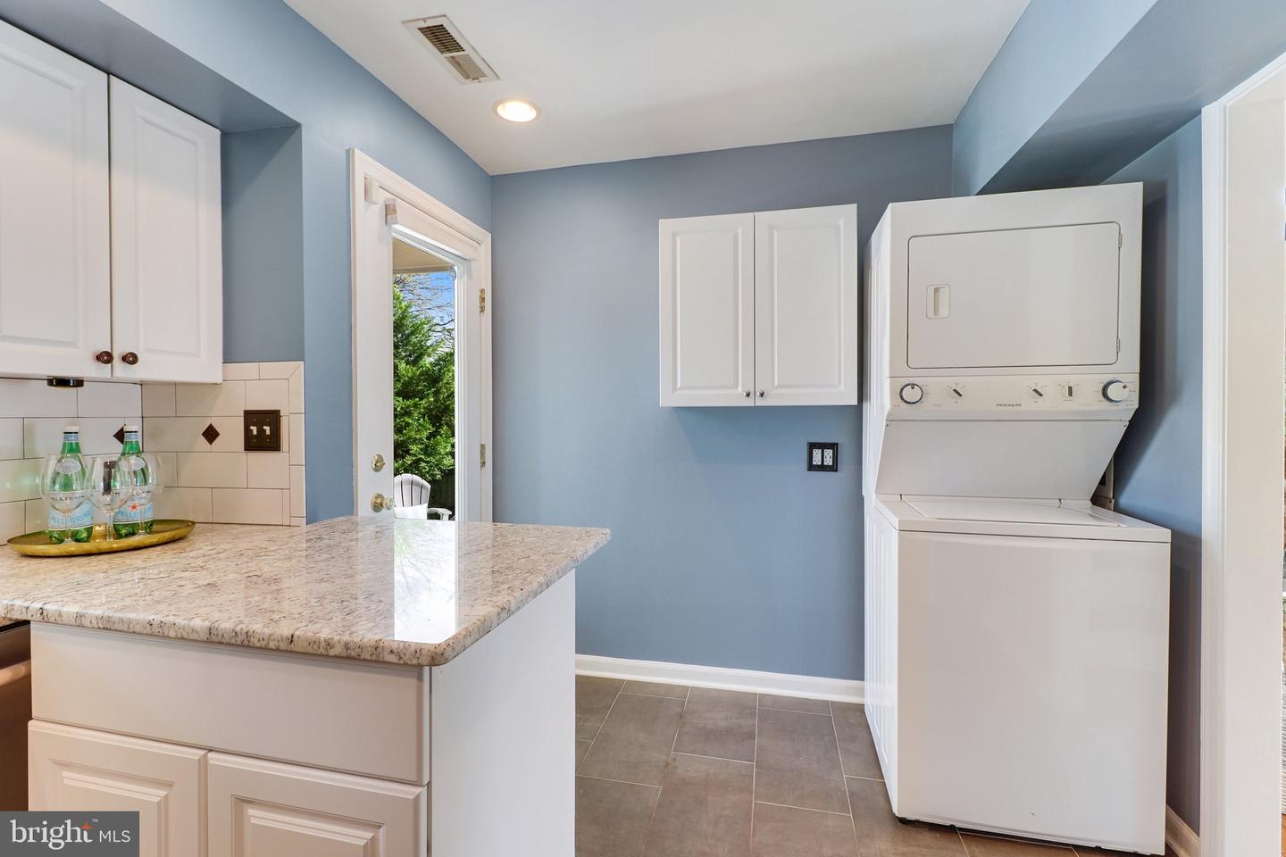 Laundry room with stacked washer and dryer, granite counter, white cabinets, and a door to the outdoors.