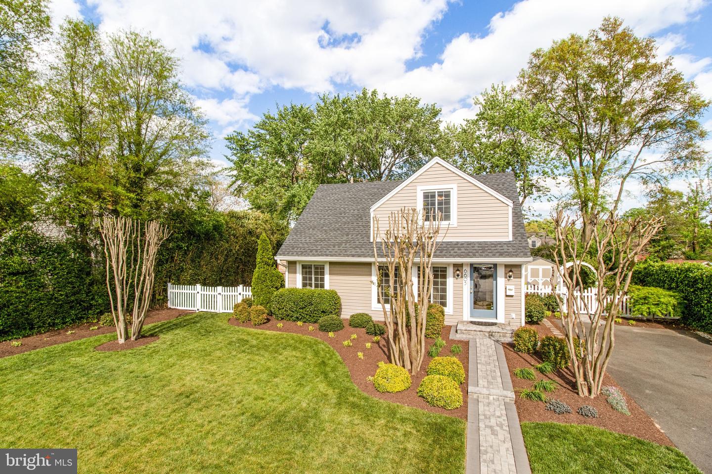 Beige two-story home with a gray roof, landscaped front yard, paved walkway, and white picket fence.