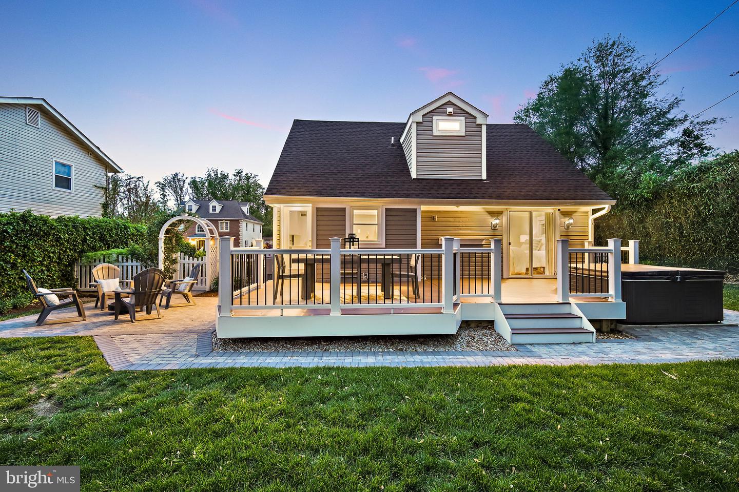 House rear with an illuminated deck, patio seating, and hot tub at dusk.