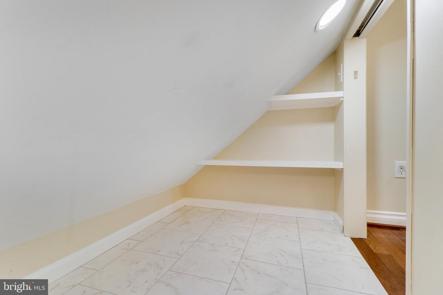 Tiled nook beneath a sloped ceiling with two built-in shelves and a doorway to hardwood flooring.