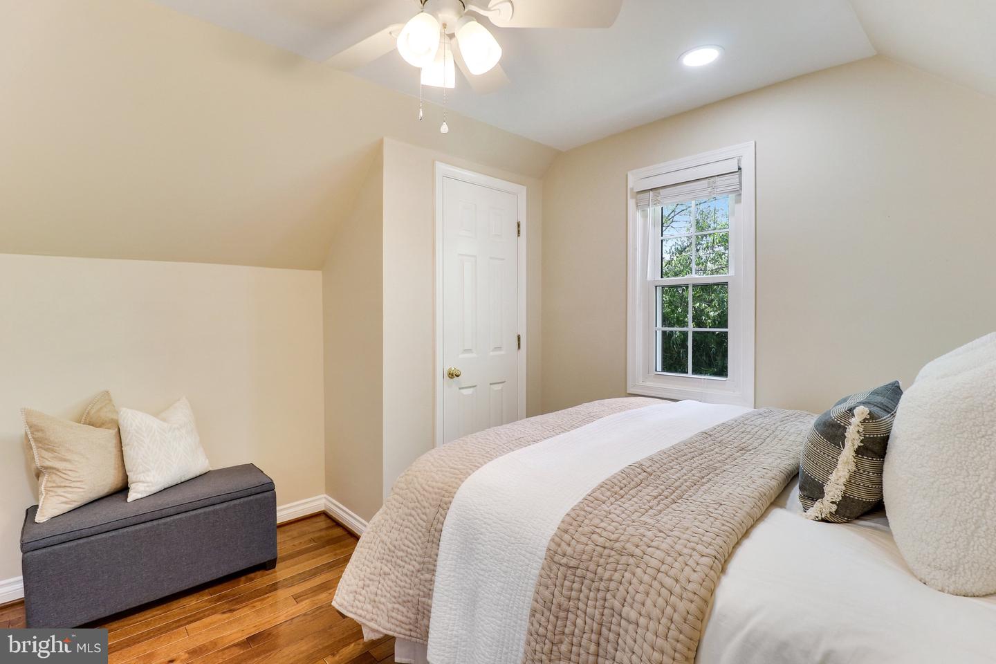 Bedroom with a beige quilted bed, hardwood floors, and a window overlooking greenery.