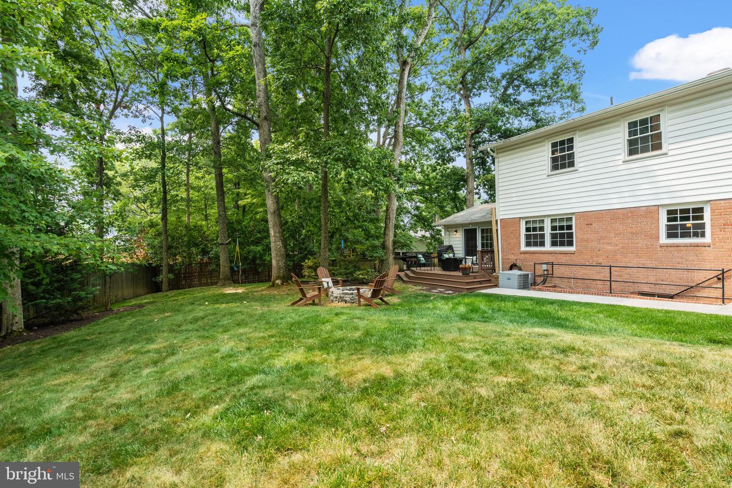 Home’s backyard with a spacious lawn, wooded surroundings, deck, and fire pit seating.