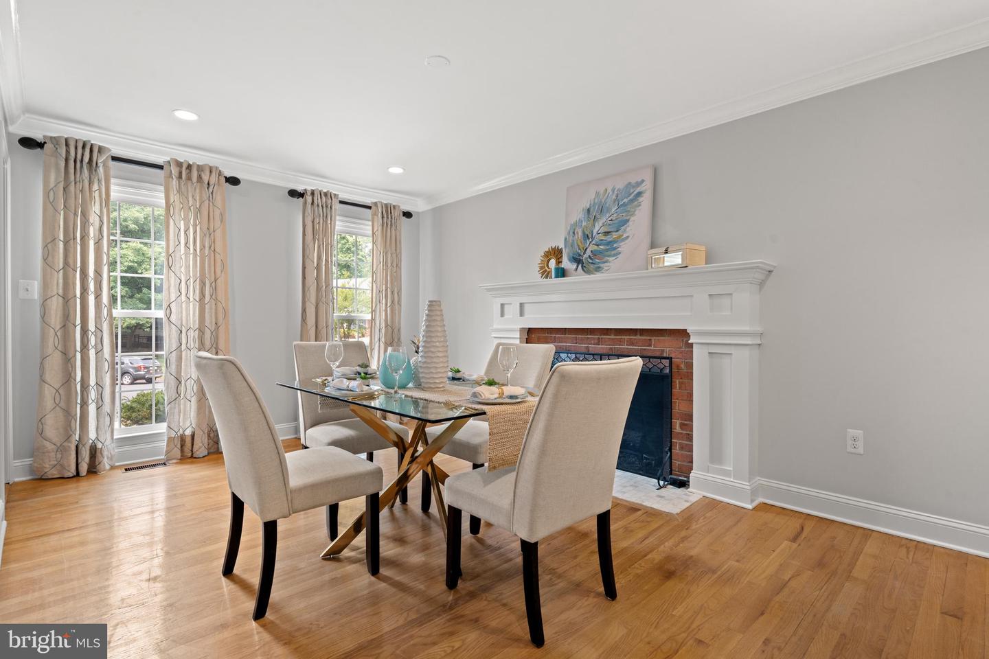 Staged dining room with a glass table, hardwood floors, and a brick fireplace with a white mantel.