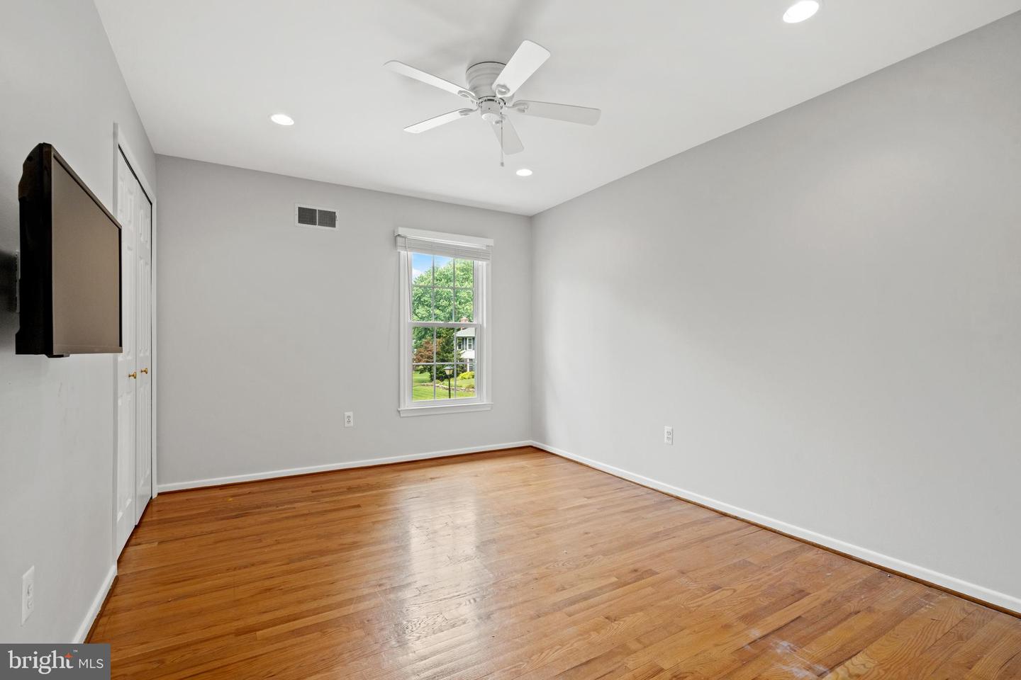 Empty bedroom with polished hardwood floors, a ceiling fan, and a window overlooking greenery.