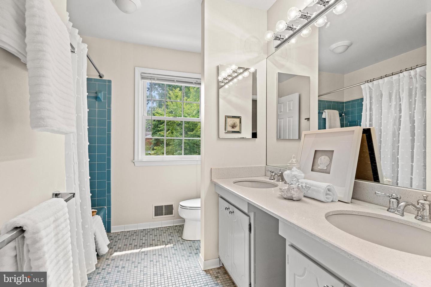 Bathroom with a double-sink vanity, blue shower tile, and a window overlooking trees.
