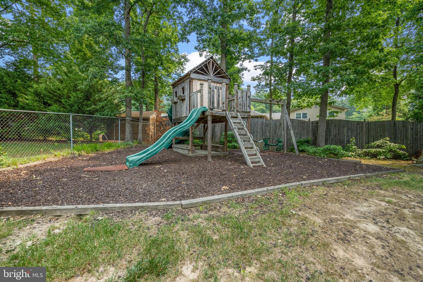 Backyard playset with green slides and ladder on a mulched area beneath mature trees.