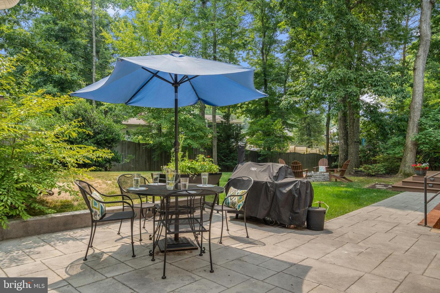 Stone patio with umbrella-covered dining set, grill, lawn, and wooded fenced yard.