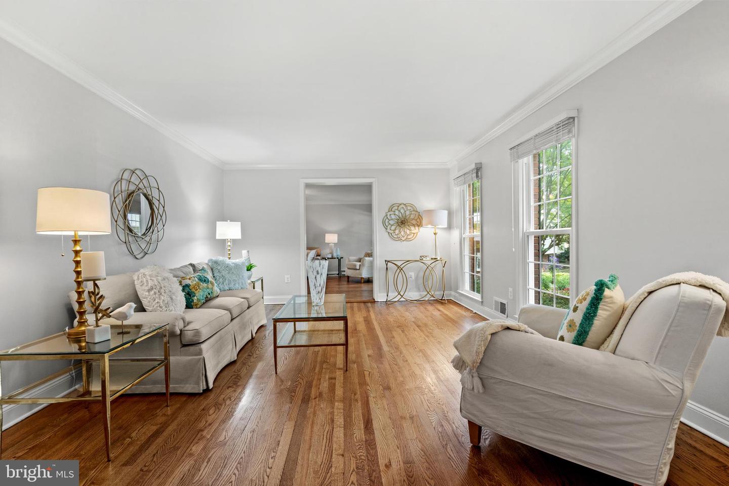 Staged living room with hardwood floors, neutral seating, and tall windows overlooking greenery.