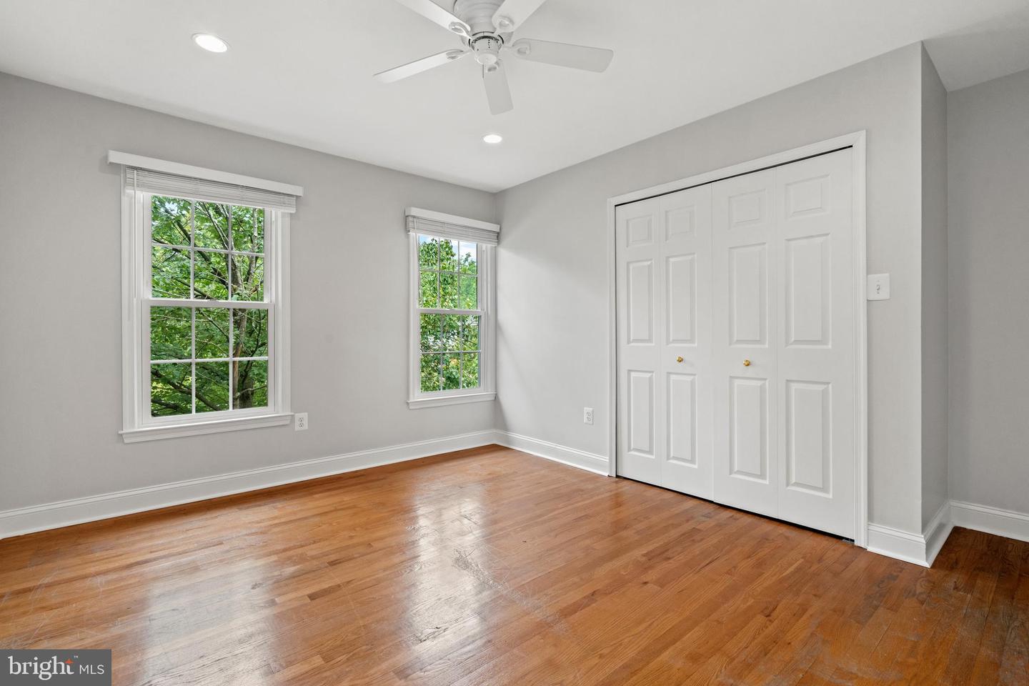 Empty bedroom with hardwood floors, two windows, a ceiling fan, and white double closet doors.