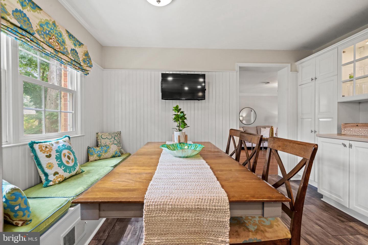 Dining nook with a wooden table, green window bench, patterned shade, and white built-in cabinets.