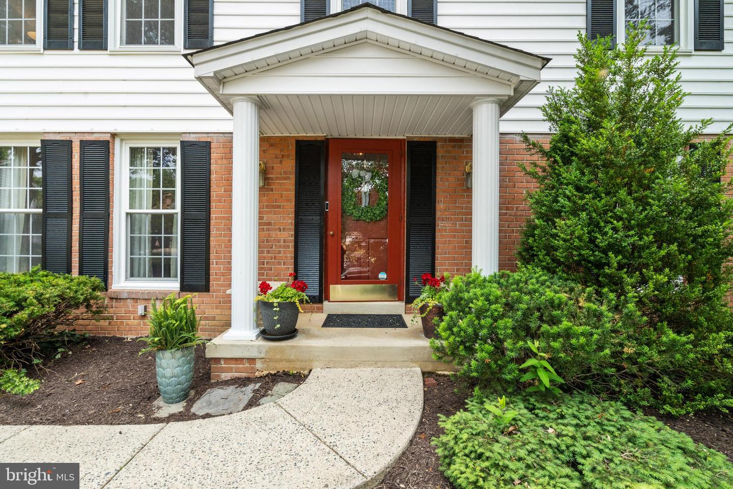 Brick home entrance with a red wreath-adorned door, white porch columns, potted flowers, and landscaped shrubs.