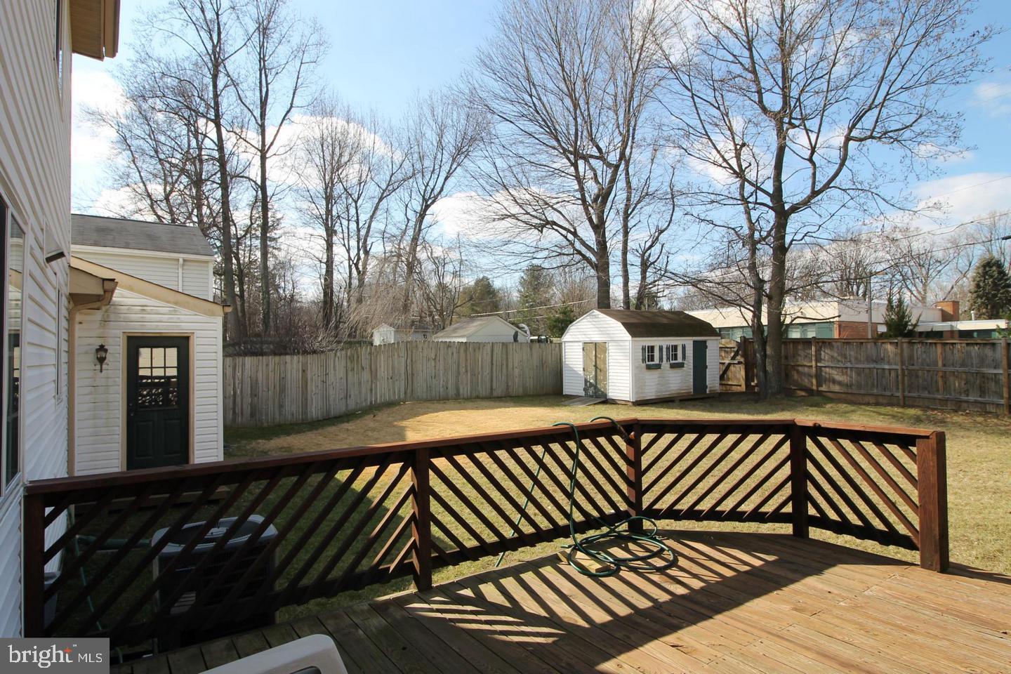 Fenced backyard with a wooden deck, lawn, and white storage shed beneath leafless trees.