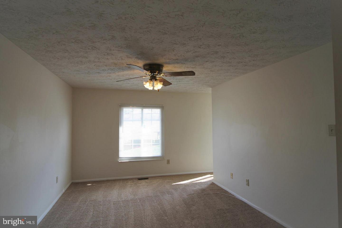Empty carpeted room with a ceiling fan and a sunlit window with blinds.