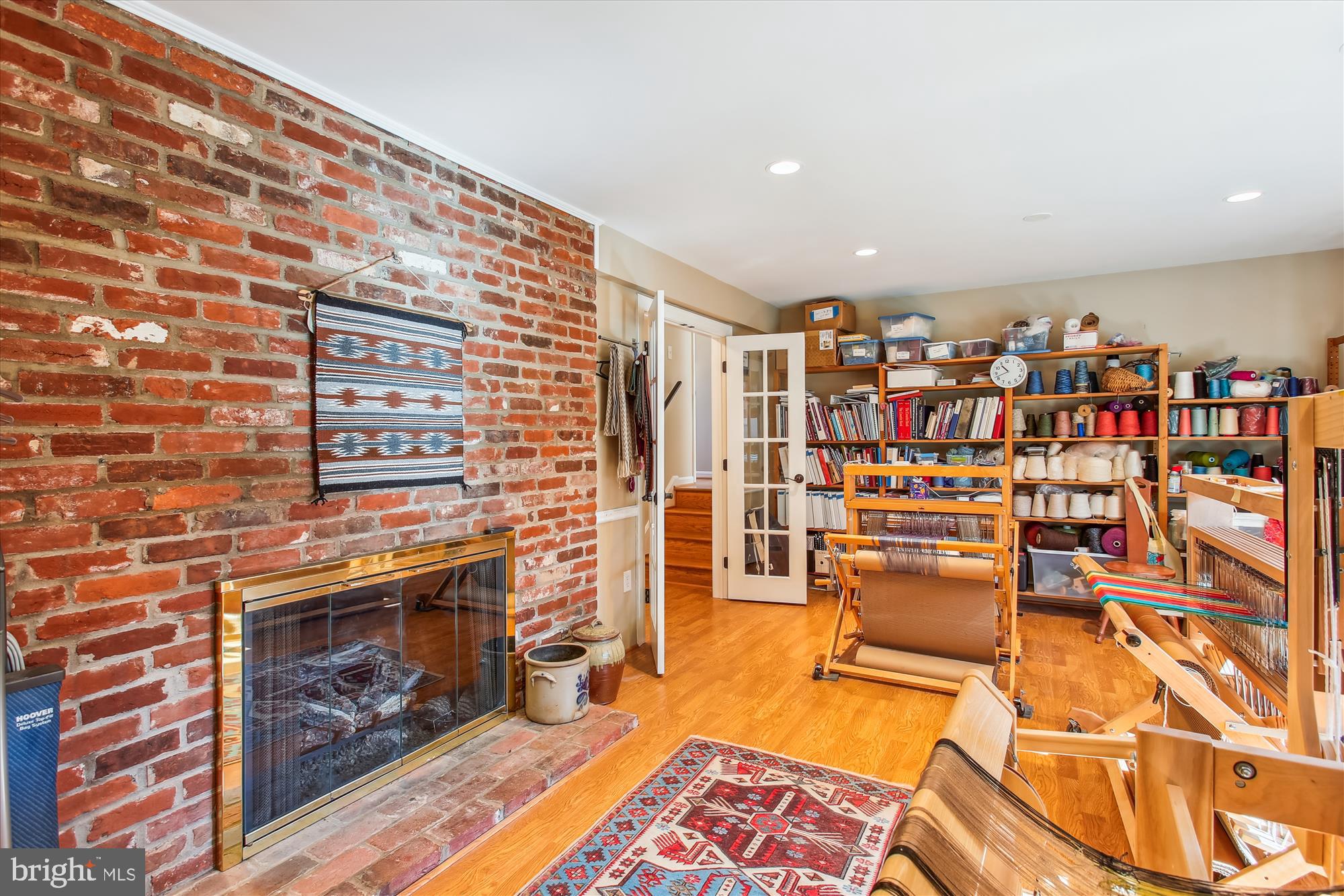 Weaving studio with looms and yarn shelves beside a brick fireplace.