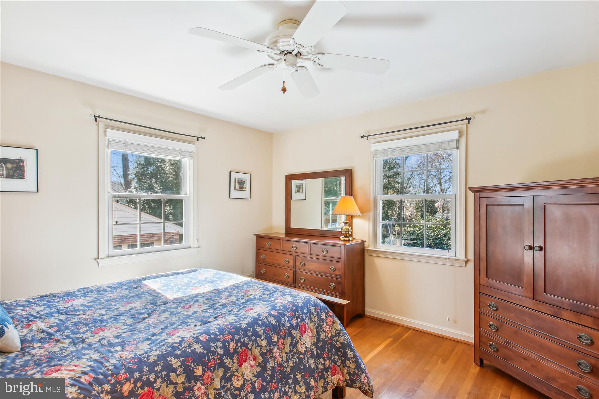 Bedroom with a blue floral bedspread, hardwood floors, and dark wood dressers between two windows.