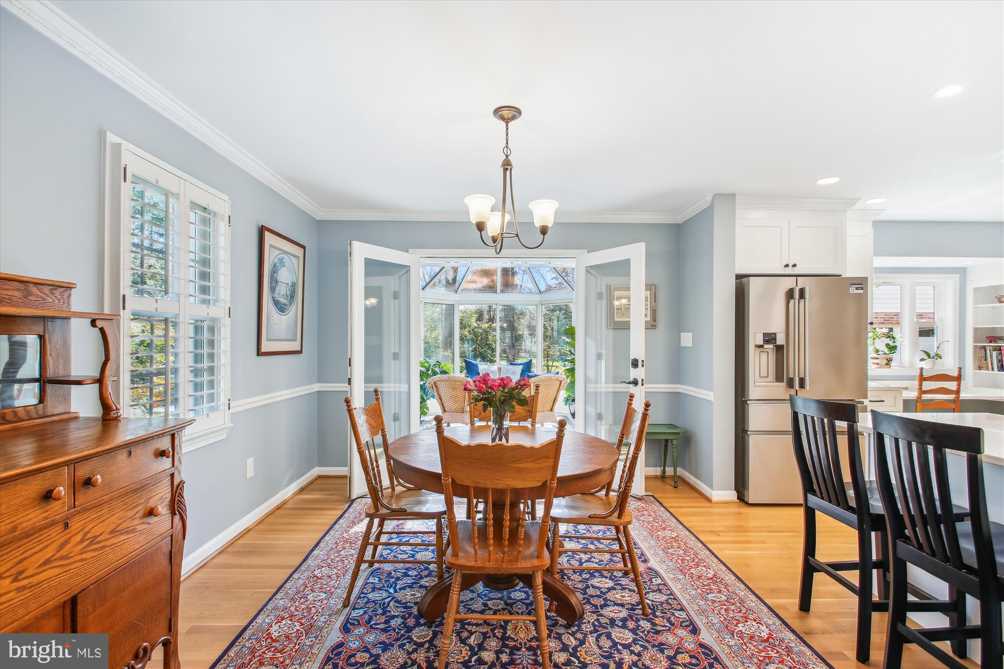 Dining room with a round wooden table, patterned rug, plantation shutters, and glass doors to a sunroom.