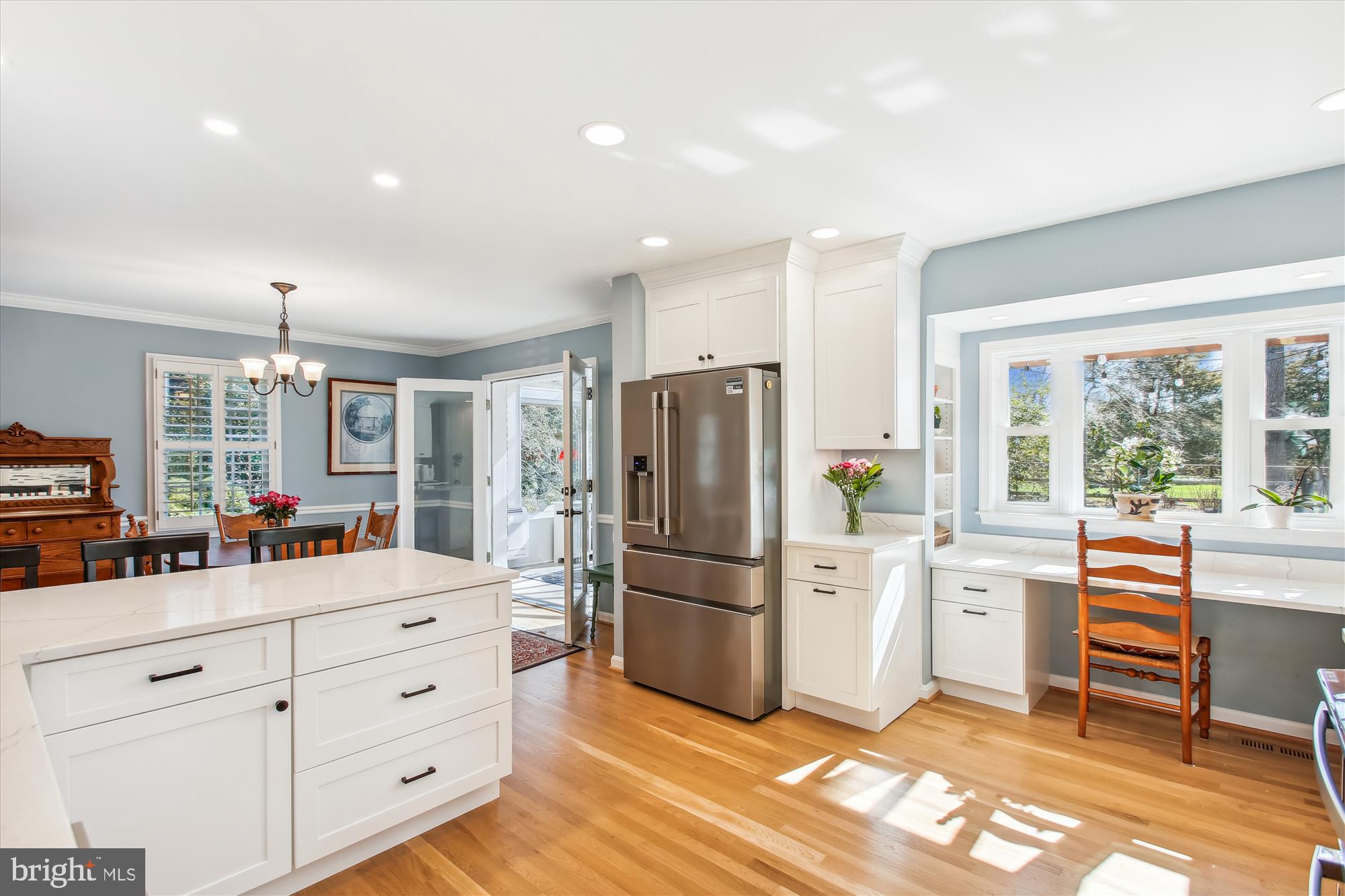 Bright kitchen with a white island, stainless steel refrigerator, hardwood floors, and a sunny window-side counter.