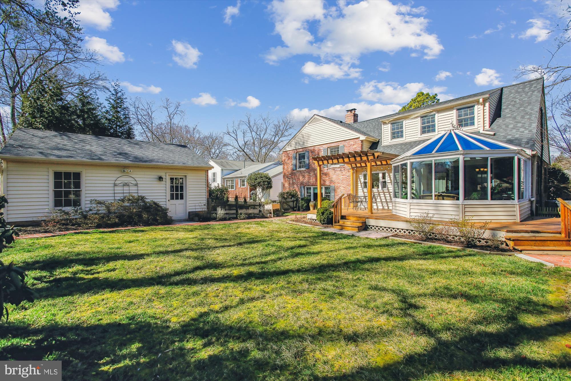 Rear exterior of a brick-and-siding home with a glass sunroom, deck, pergola, and broad lawn.
