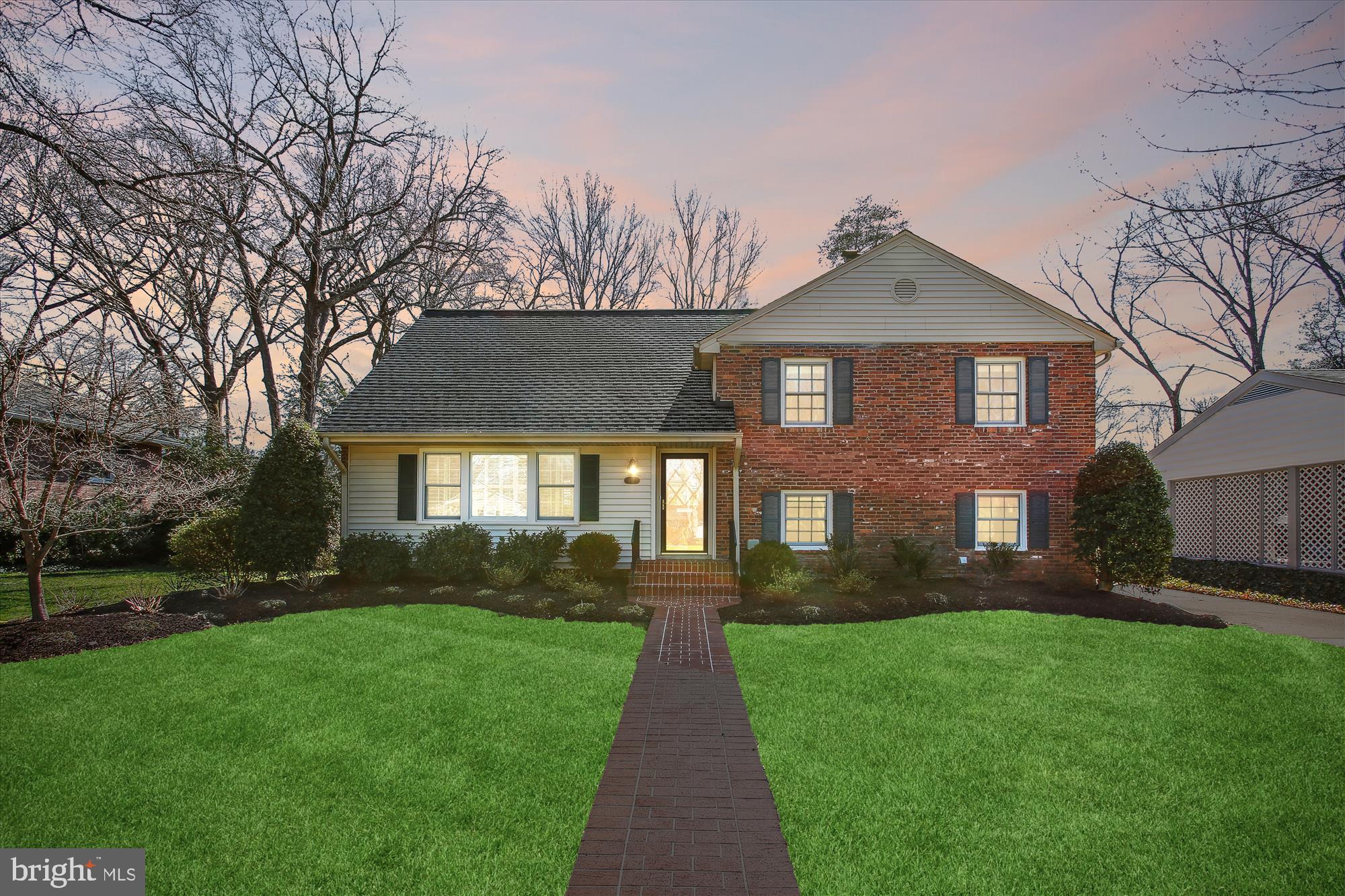 Two-story brick and gray-sided house with a brick walkway, green lawn, and illuminated windows at dusk.