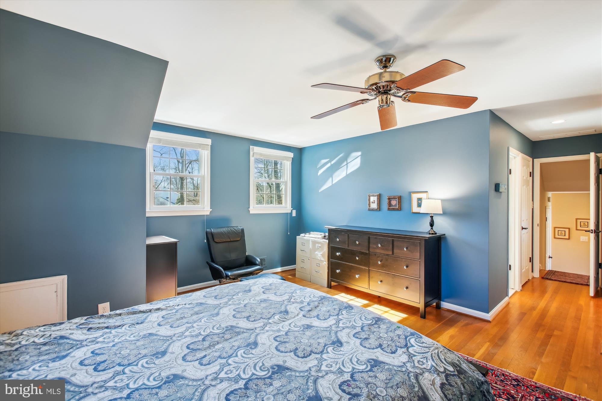 Furnished blue bedroom with patterned bedding, hardwood floors, two windows, and a ceiling fan.