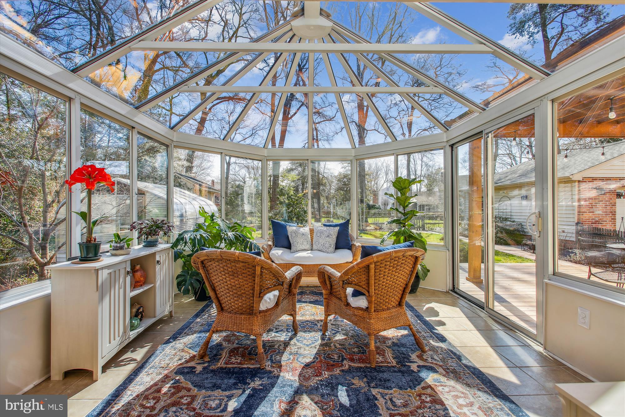 Sunroom with a peaked glass roof, wicker seating, and potted plants overlooking the backyard.