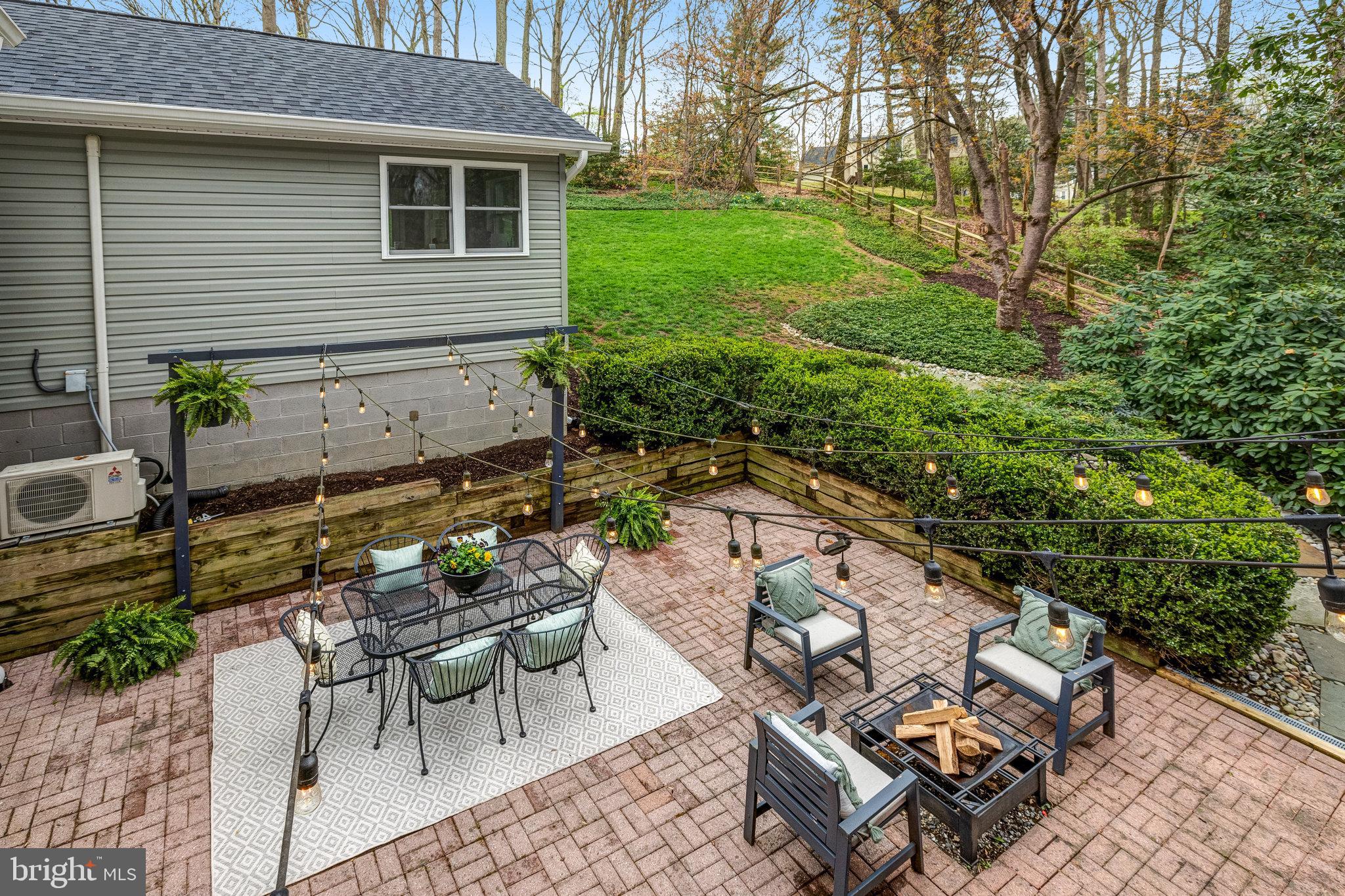 Brick patio with lounge seating, a fire pit, and string lights beside a landscaped lawn.