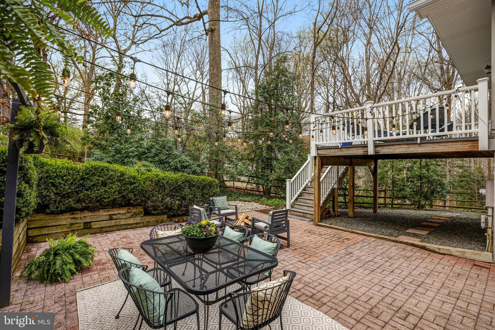Brick-paver patio with outdoor dining furniture, string lights, and stairs to a raised deck.