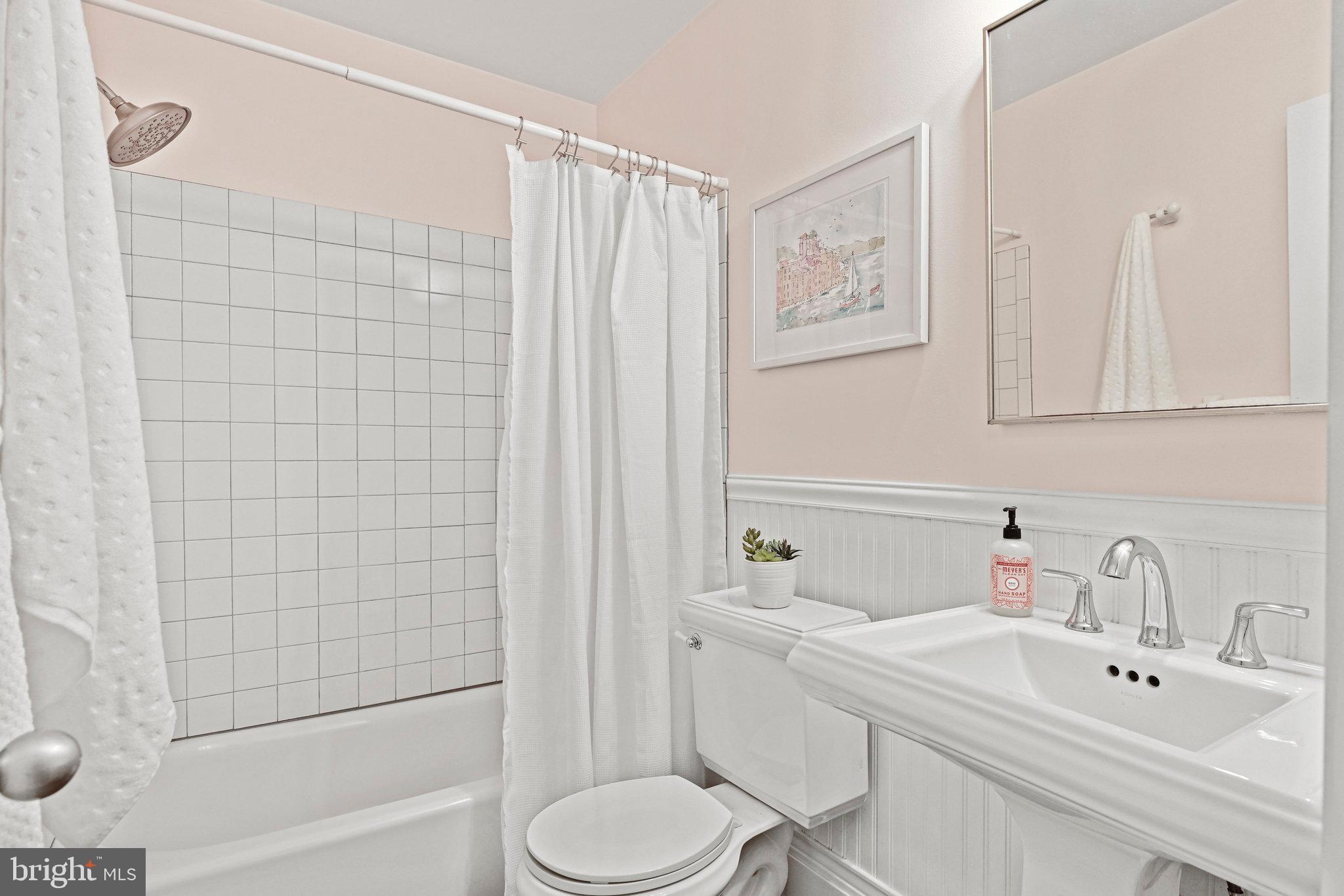 White bathroom with tiled tub and shower, pedestal sink, and framed artwork.