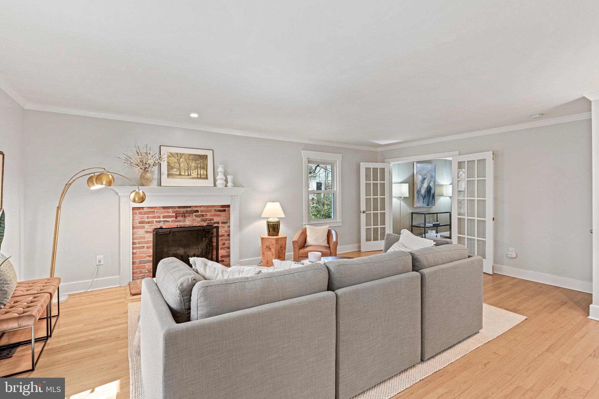 Staged living room with a gray sectional, red-brick fireplace, hardwood floors, and open French doors.