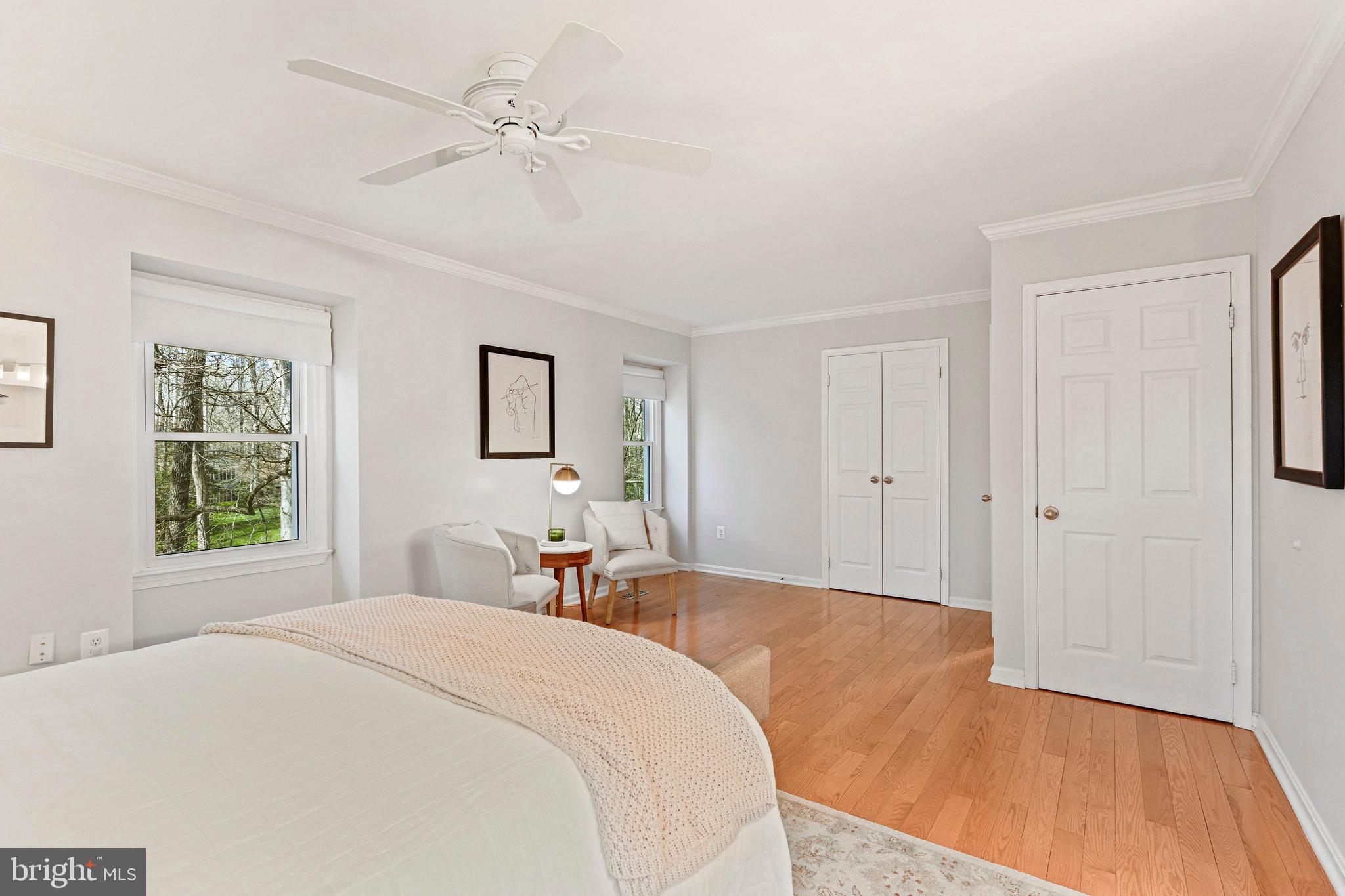 Furnished bedroom with hardwood floors, a ceiling fan, and a sitting area beside the windows.