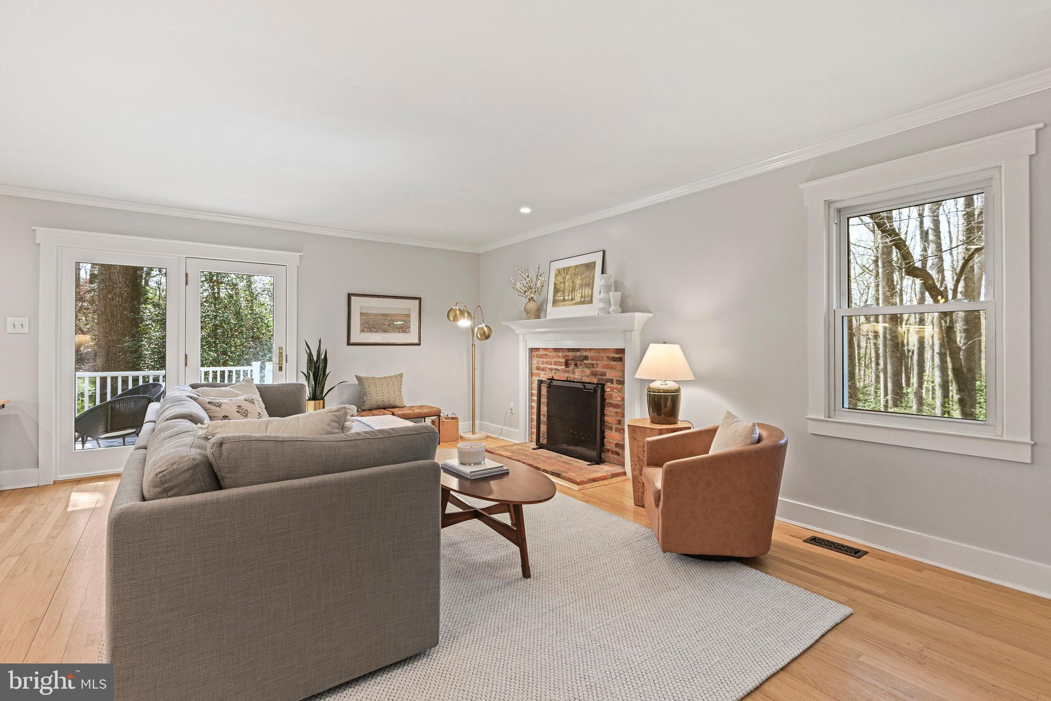 Staged living room with brick fireplace, gray sectional, and windows overlooking trees.