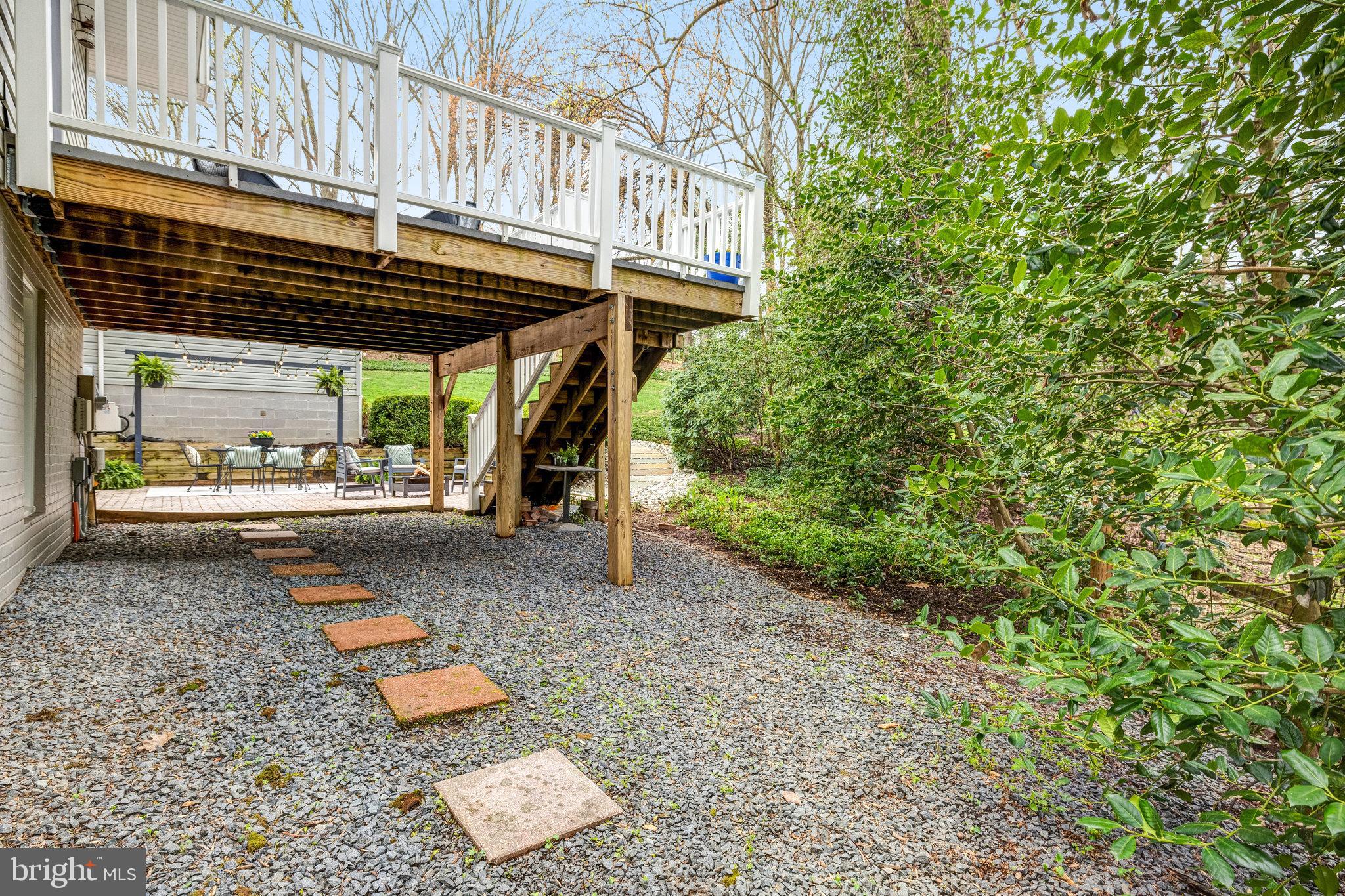 Backyard patio beneath a raised deck, with a gravel stepping-stone path and leafy landscaping.