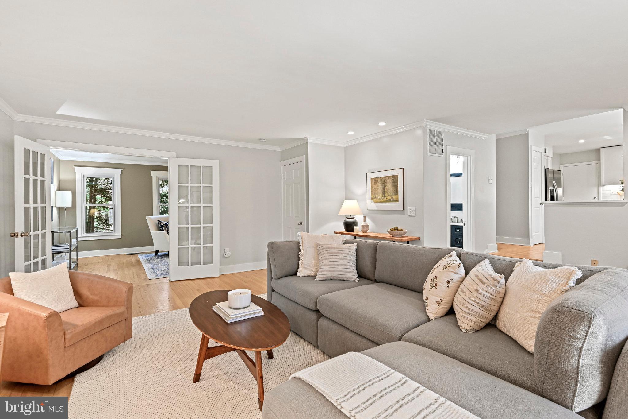 Staged living room with a gray sectional, tan armchair, and French doors to an adjoining sitting room.