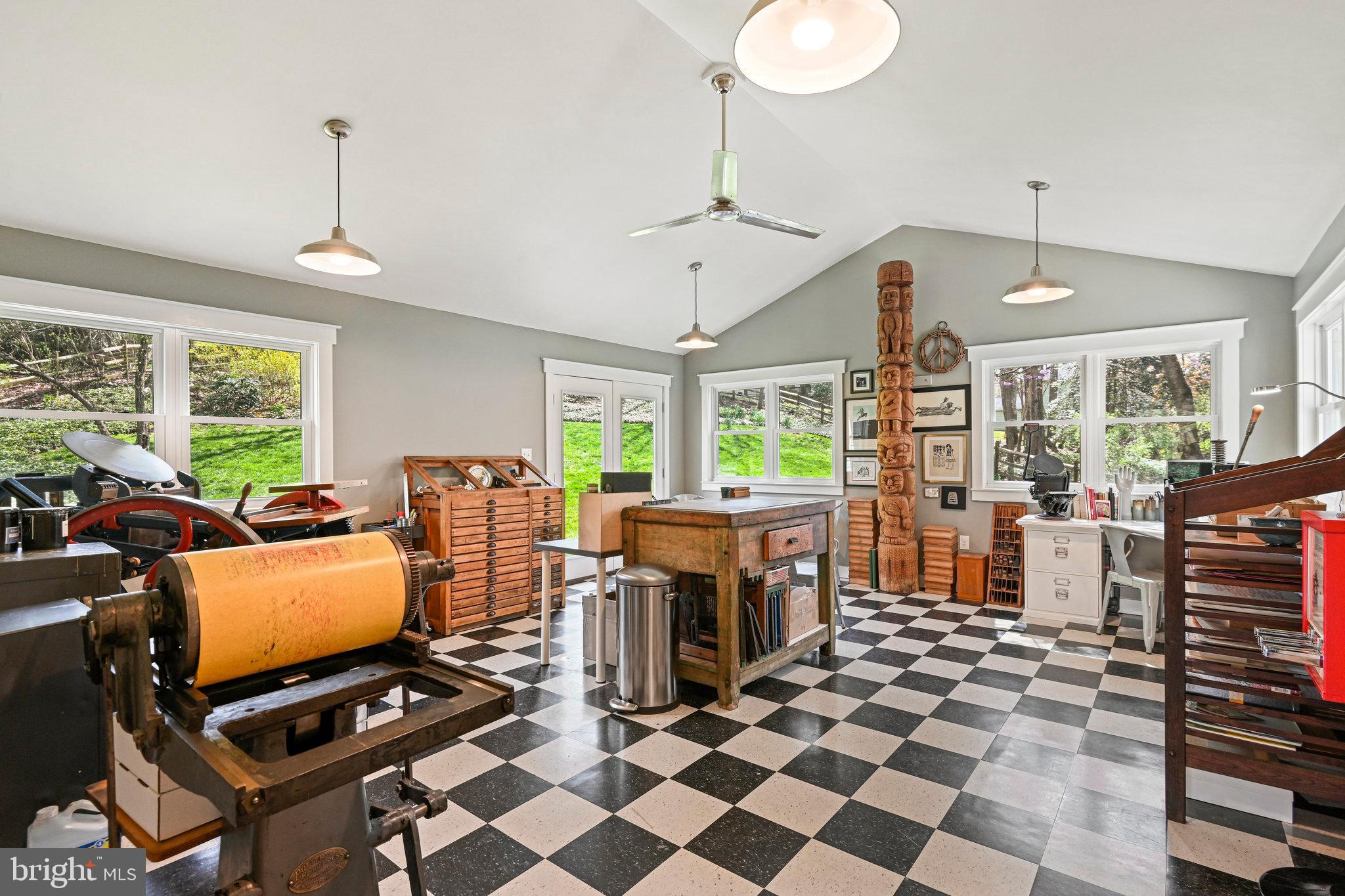 Workshop with printing equipment, wooden worktables, checkerboard floor, and large windows.