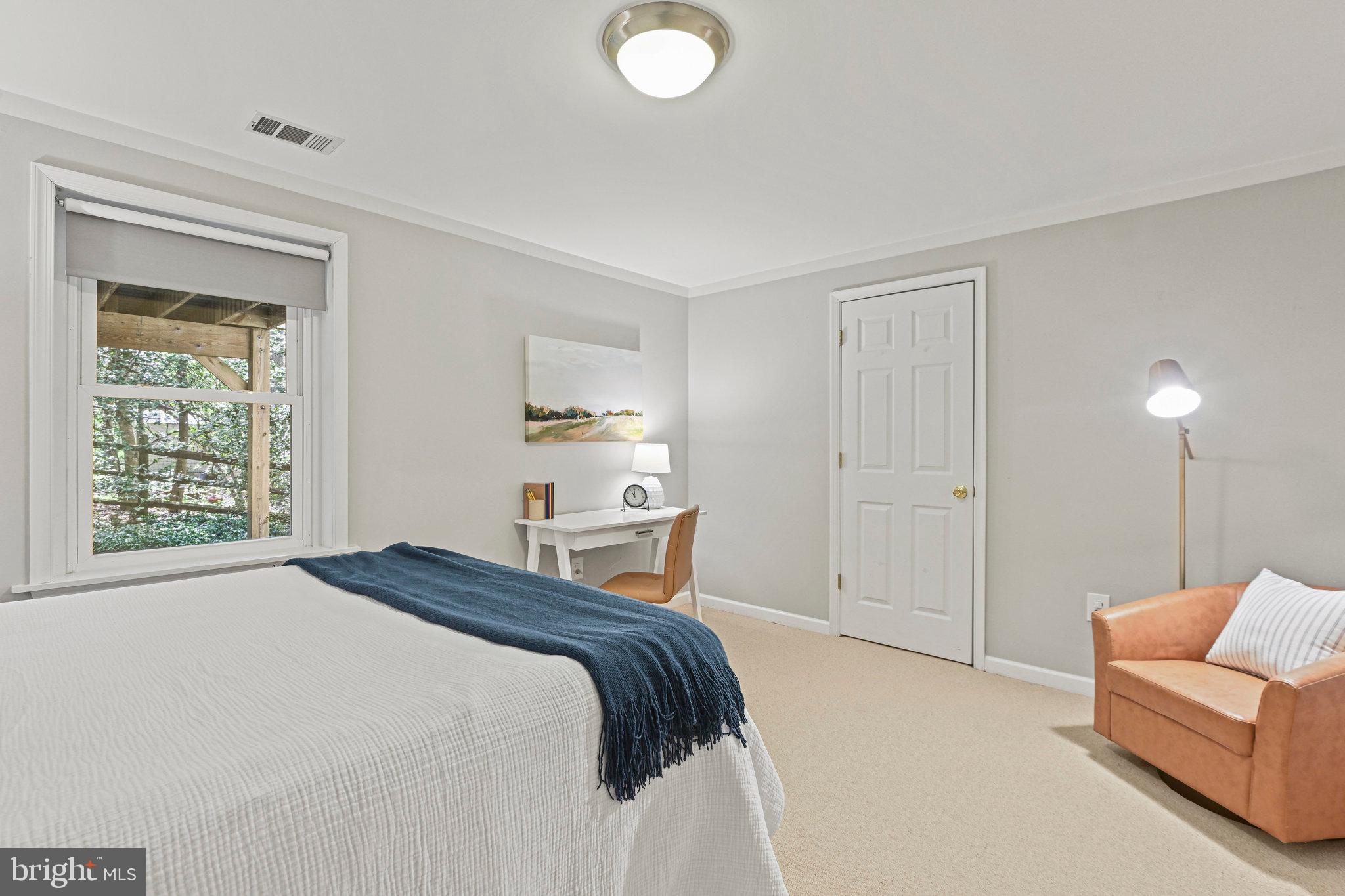Bedroom with a white bed, navy throw, desk, and tan armchair beside a window.